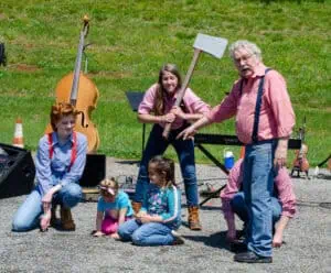 Three Jack Tale Players in red and blue gingham shirts and suspenders in front of musical instruments outdoors, with one holding a fake axe and Rex Stephenson pointing to two small girls in a Jack Tale show