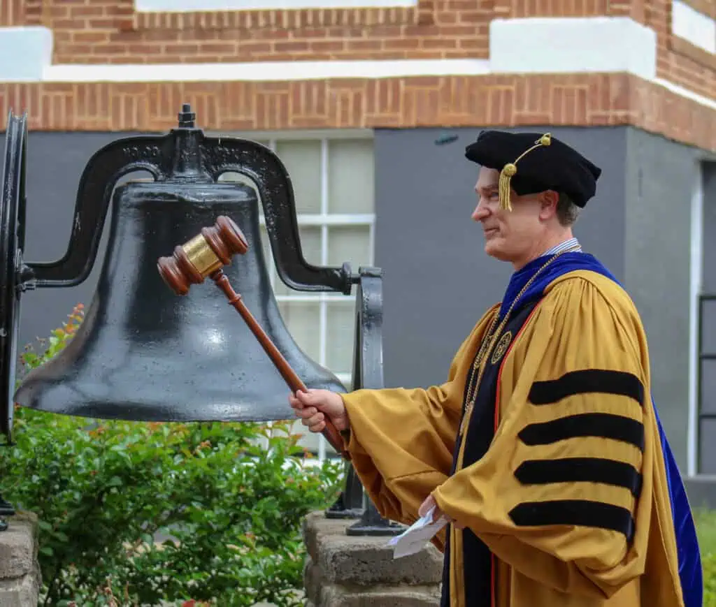 Ferrum College President David Johns in gold and black academic robes ringing the Schoolfield Hall bell for the first time at graduation in 2019
