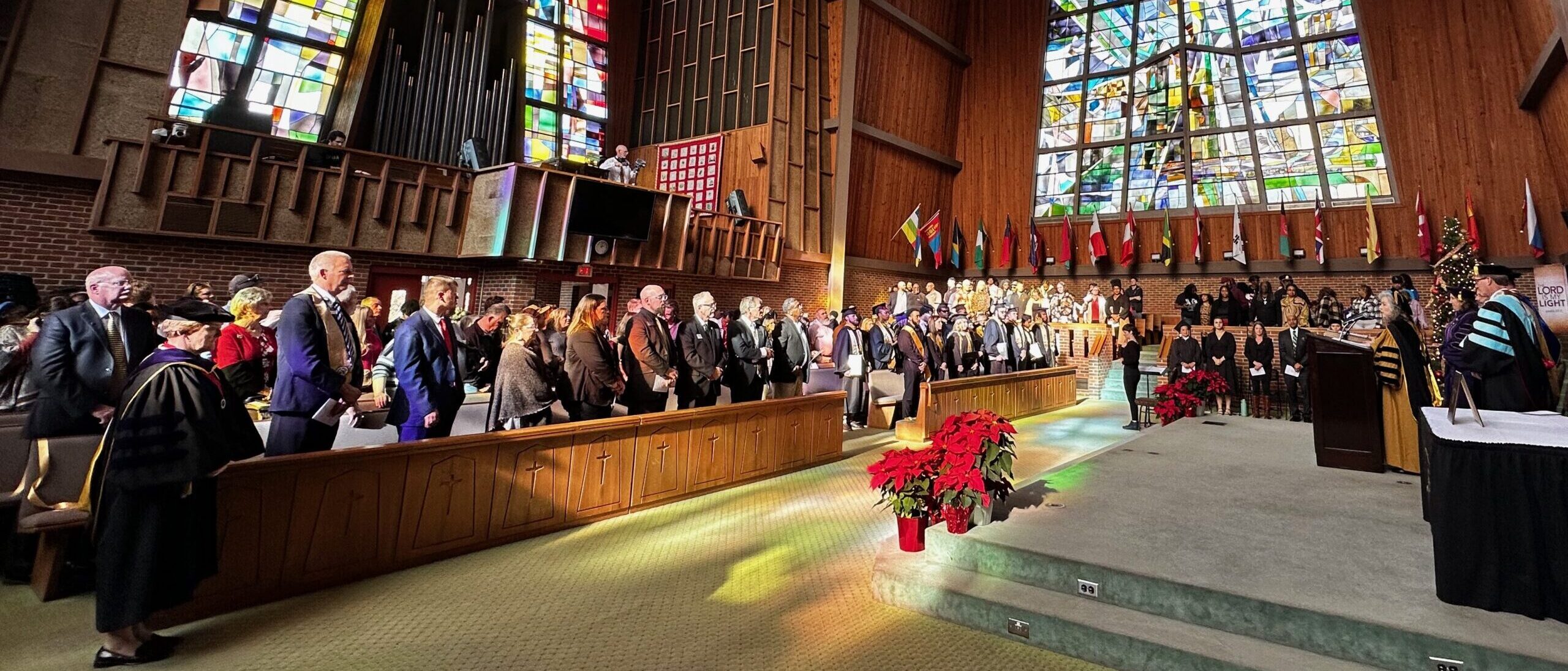 Photograph taken in Vaughn Chapel of standing audience members attending the fall commencement, including graduates, poinsettias, flags, the platform party, and stained glass windows