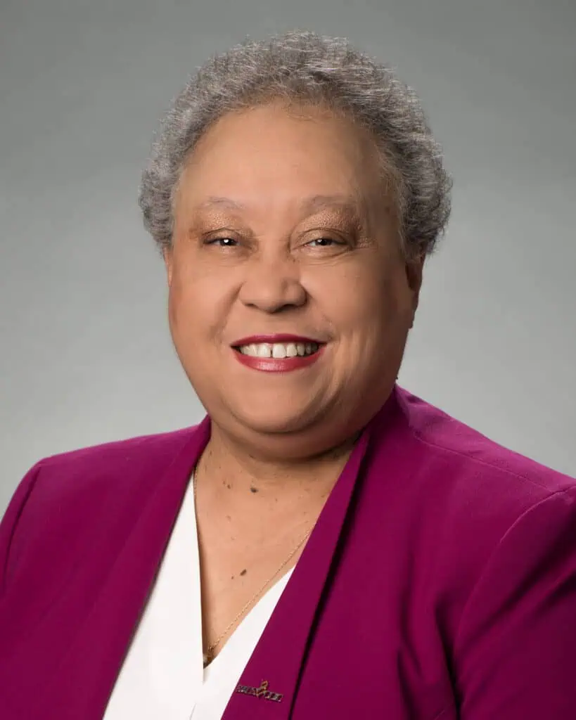 Head shot of Dr. Belle Wheelan on a grey background, with very short hair, a purple jacket, and white blouse.