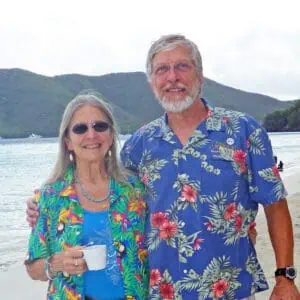 Bob and Carolyn Thomas in front of a lake and hills, both in flowered shirts, with Carolyn in sunglasses holding a cup