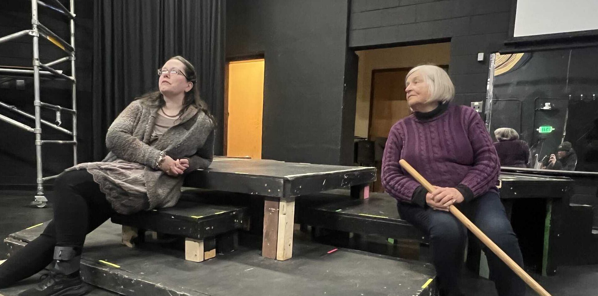 Two actors in dark clothing rehearsing in a black-box theatre, sitting on low platforms