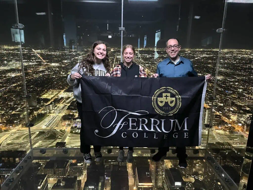 Ed Hally and 2 honors students holding Ferrum College banner in a glass big city overlook with dark night cityscape behind them