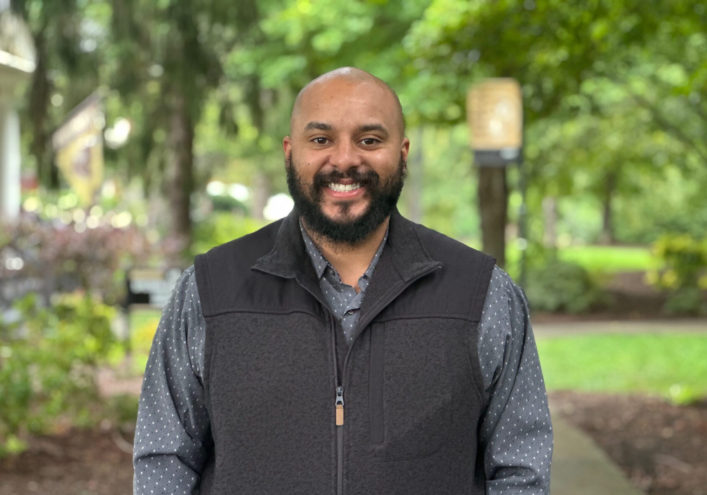 Photograph of Sam Martin wearing a gray zip-up vest over a gray shirt taken outside at Ferrum College on a pretty summer day.