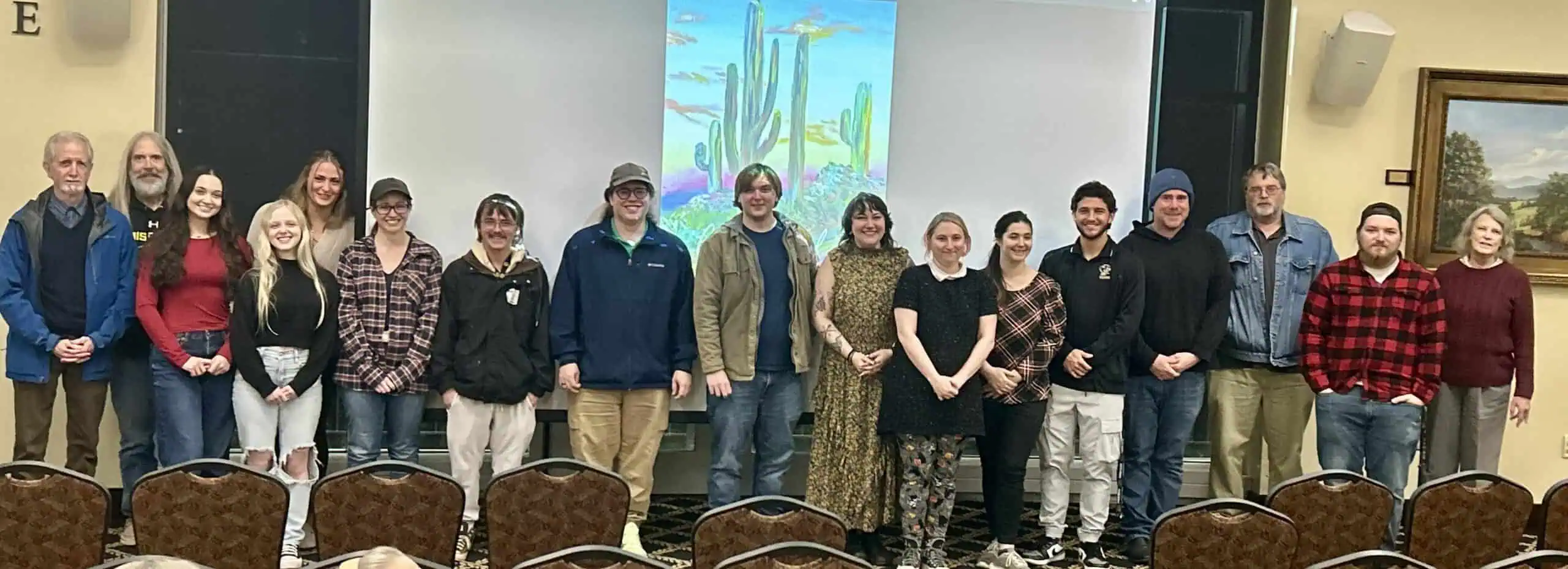 Contributors to fall 2025 Chrysalis standing in a row in front of colorful cover art on a screen, with advisor on right.