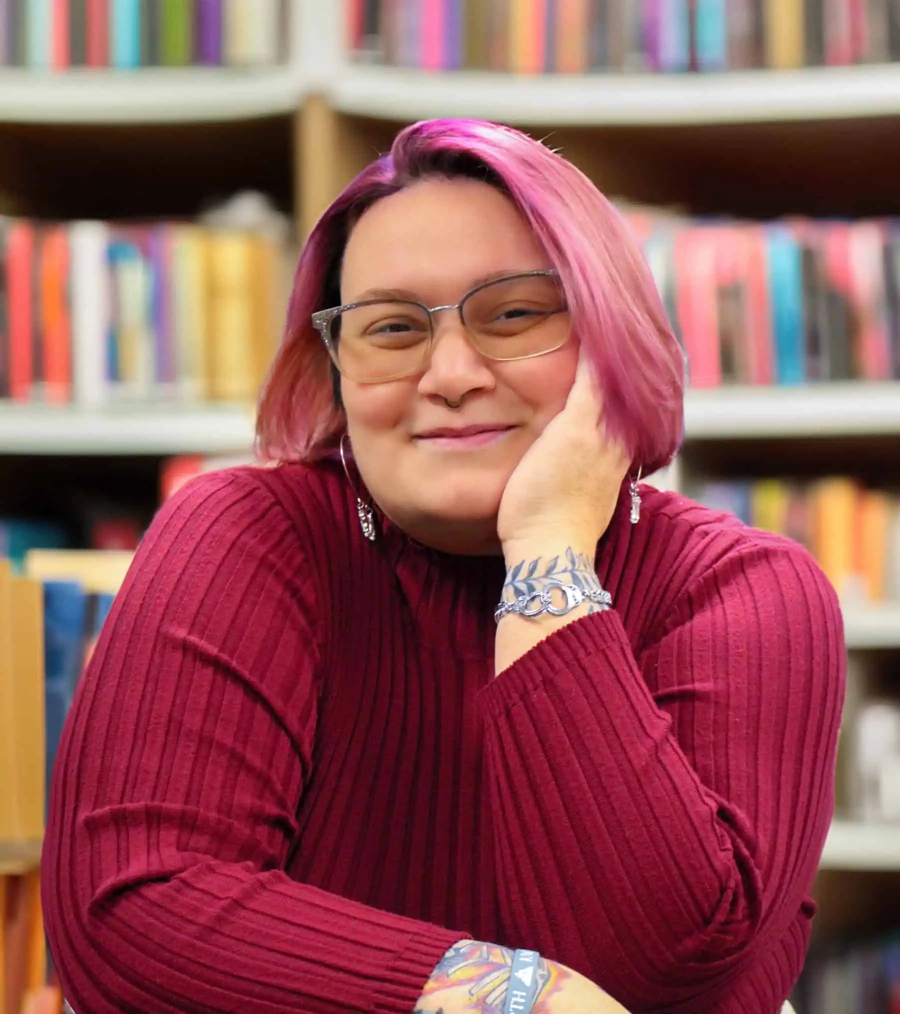 Headshot of Rachel Walton '15 with dark pink sweater and pink hair, wearing glasses in front of shelves of books