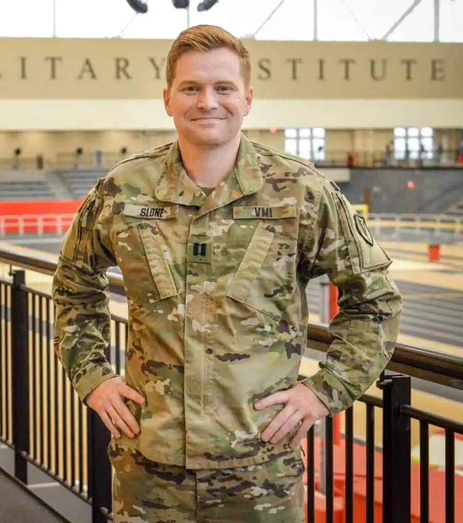 Major Isaac Slone '11 in fatigues, standing with hands on hips in front of a black balcony railing over a gym with part of the VMI name on the wall behind him.