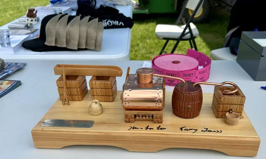 Miniature moonshine still in front of caps, shirts, and pink tickets at merch table at 2025 BRIM BBQ Cook Off event