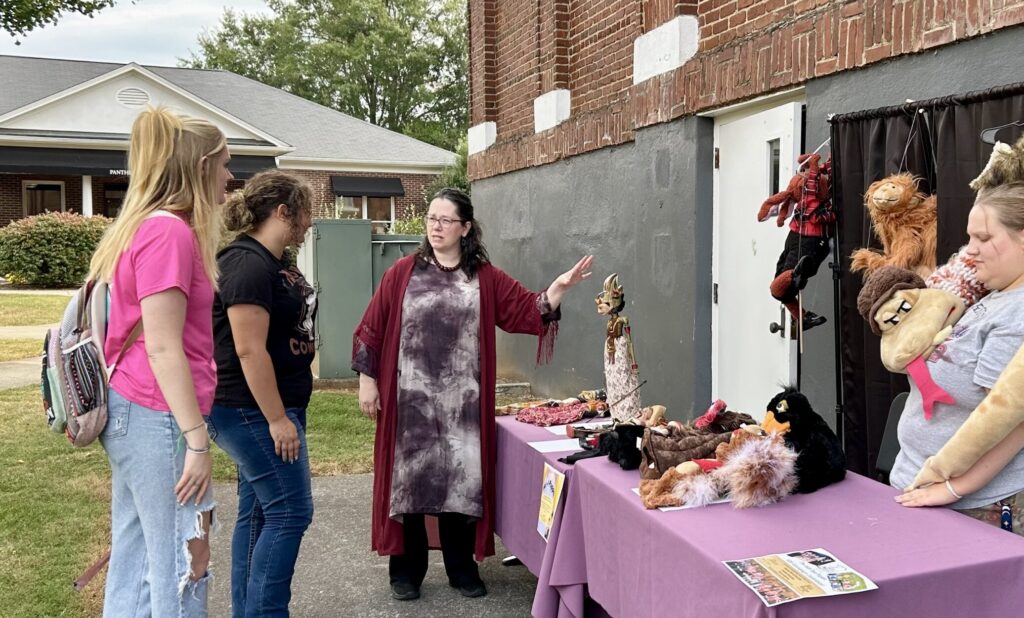 Rebecca Crocker in long robes and Hannah Dix with big snake puppet around her show long purple table display of puppets to two students, standing behind brick and cement back wall of Schoolfield Hall