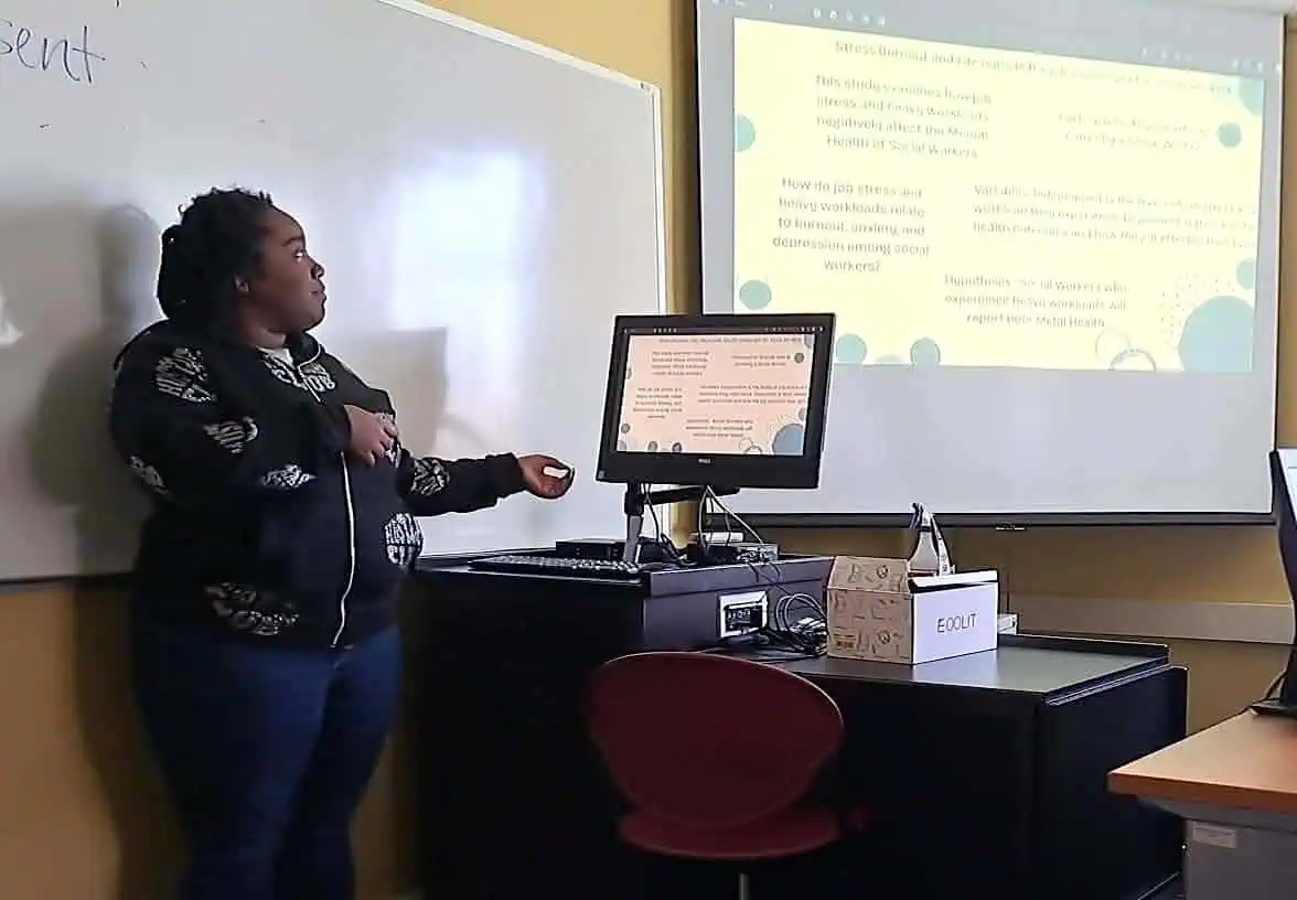 Student in a darkened classroom with a computer and screen showing her presentation slide