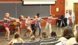 Enrichment camp kids in summer clothes and gingham Jack Tale Players shirts perform with outstretched hands with Rex Stephenson in blue shirt watching, in front of college classroom with blackboard behind them