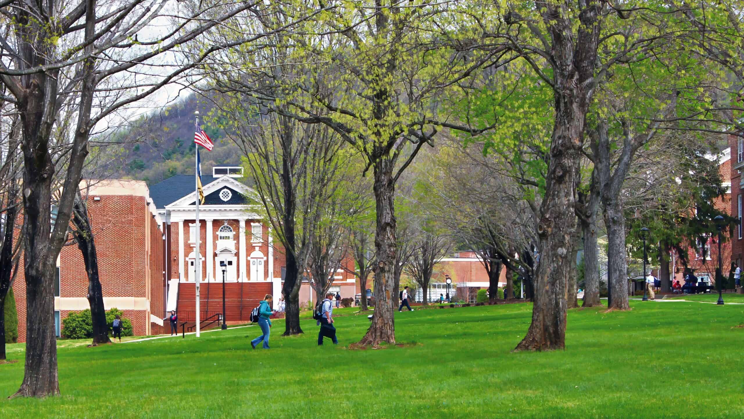 Early spring campus scene showing students walking, trees beginning to leaf out, green grass, the American flag, and brick buildings in the background