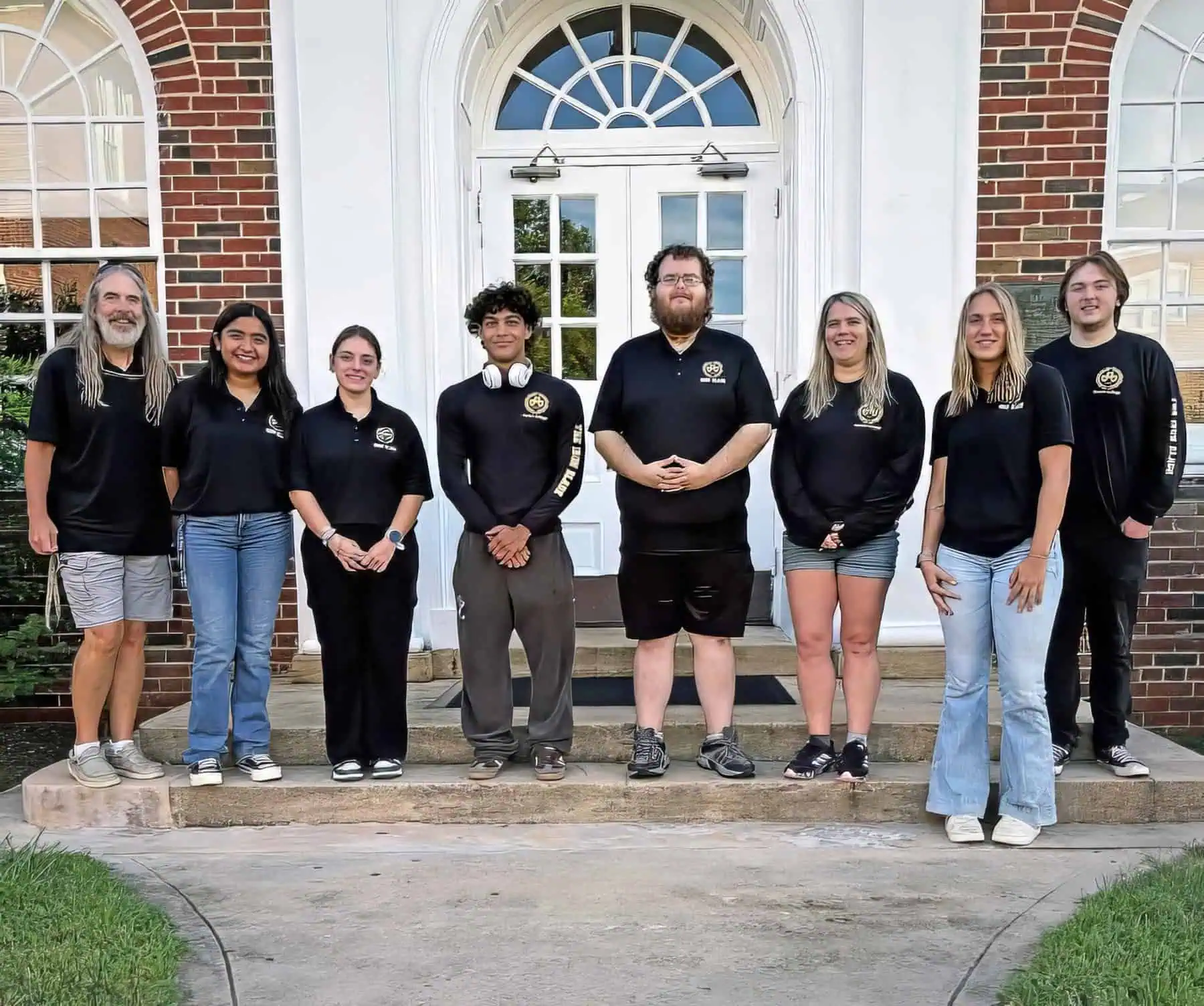 Eight members of The Iron Blade staff stand on the steps of Britt Hall wearing black Iron Blade shirts