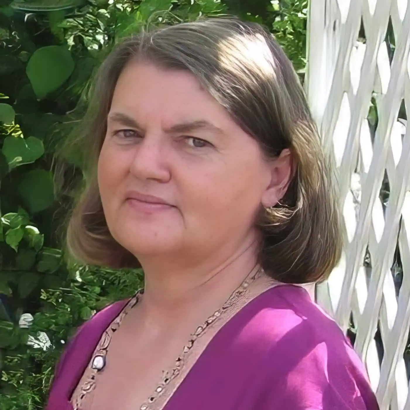 Photograph of Tina Hanlon wearing a pink dress and long necklace, standing outside with greenery and a white lattice beside her