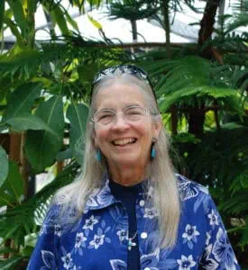 Dr. Carolyn Thomas in blue and white flowered shirt with glasses on her head standing in front of green plants