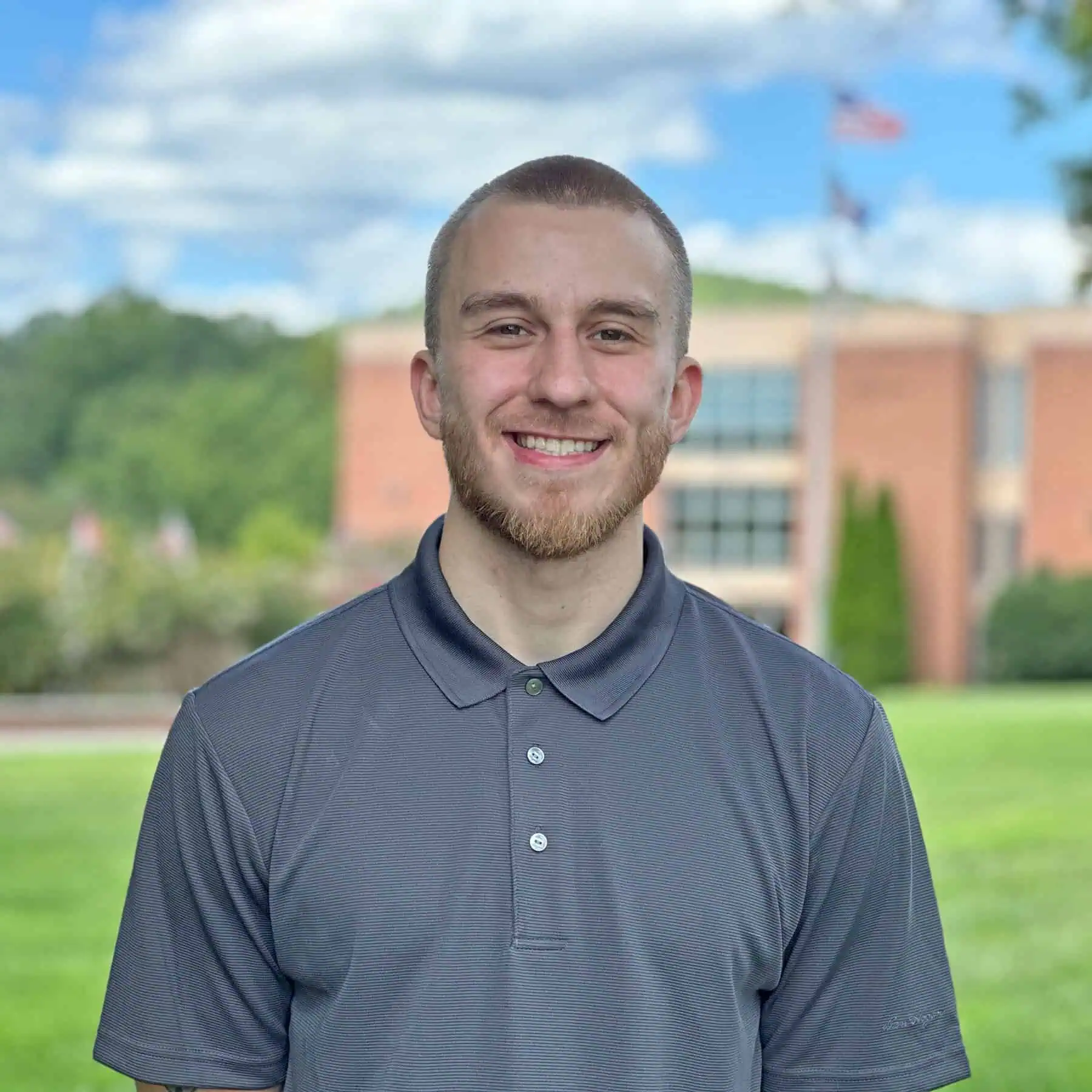 Photo of Josh Reeves standing outside on a summer day with the brick Stanley Library and a flag pole flying the US, Virginia, and Ferrum College flags behind him