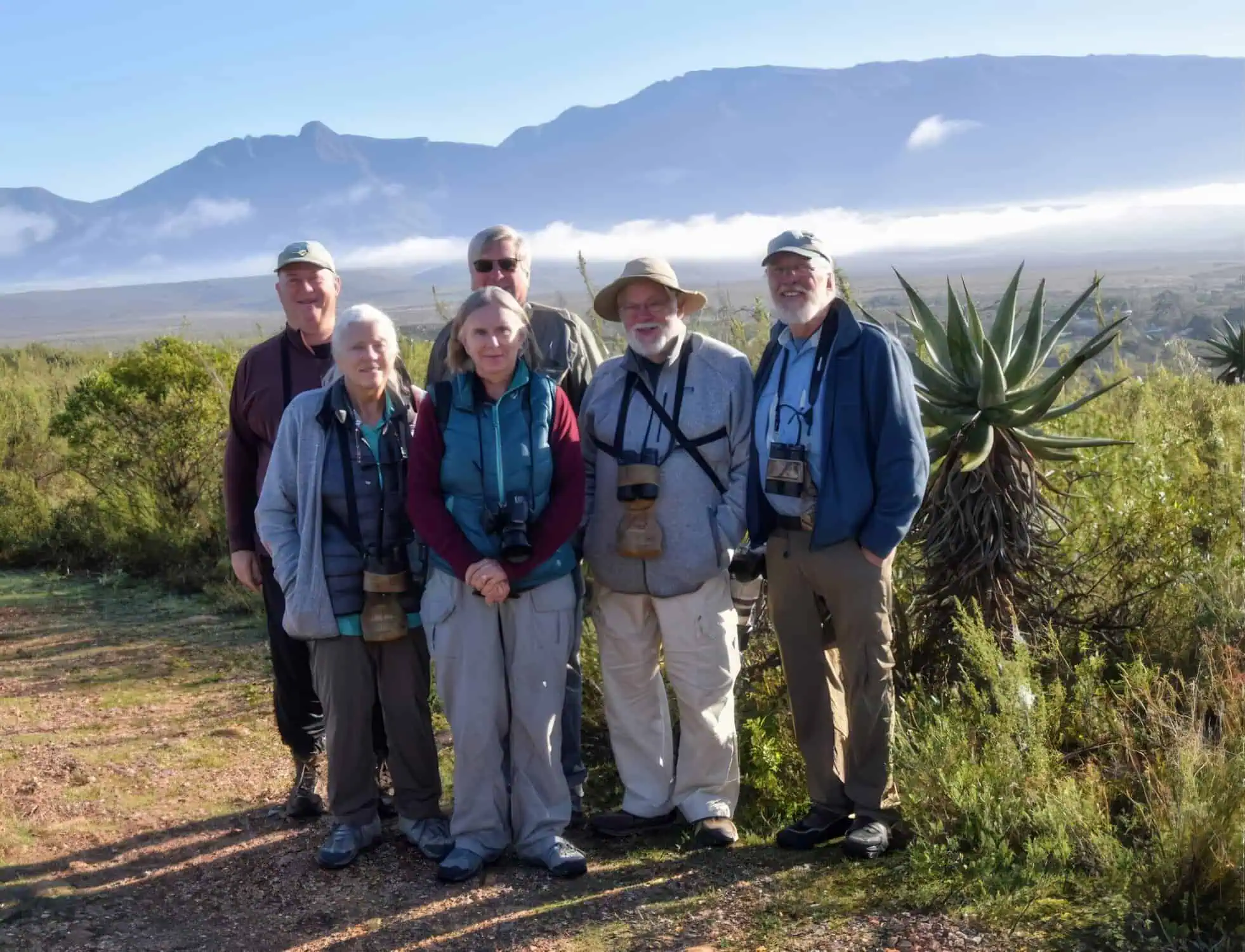 Group of six hikers wearing outdoor gear and binoculars standing on a dirt path with mountains and mist in the background.