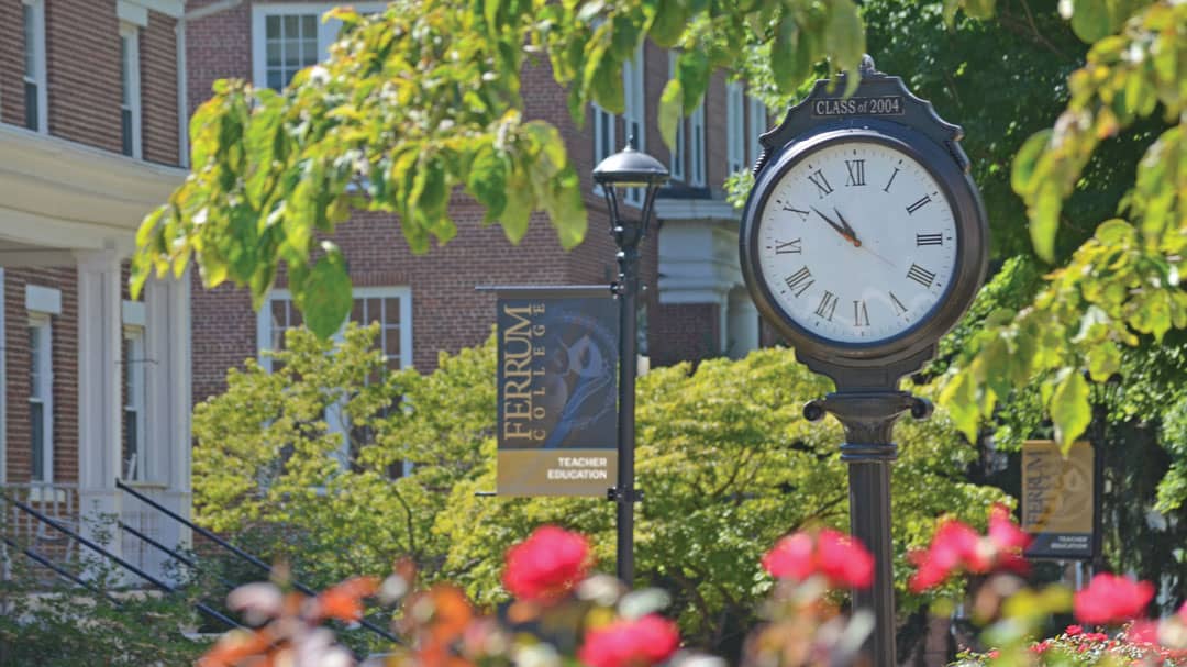 Ferrum College campus photo showing an outdoor clock, a teacher education banner, brick buildings, red flowers, and trees