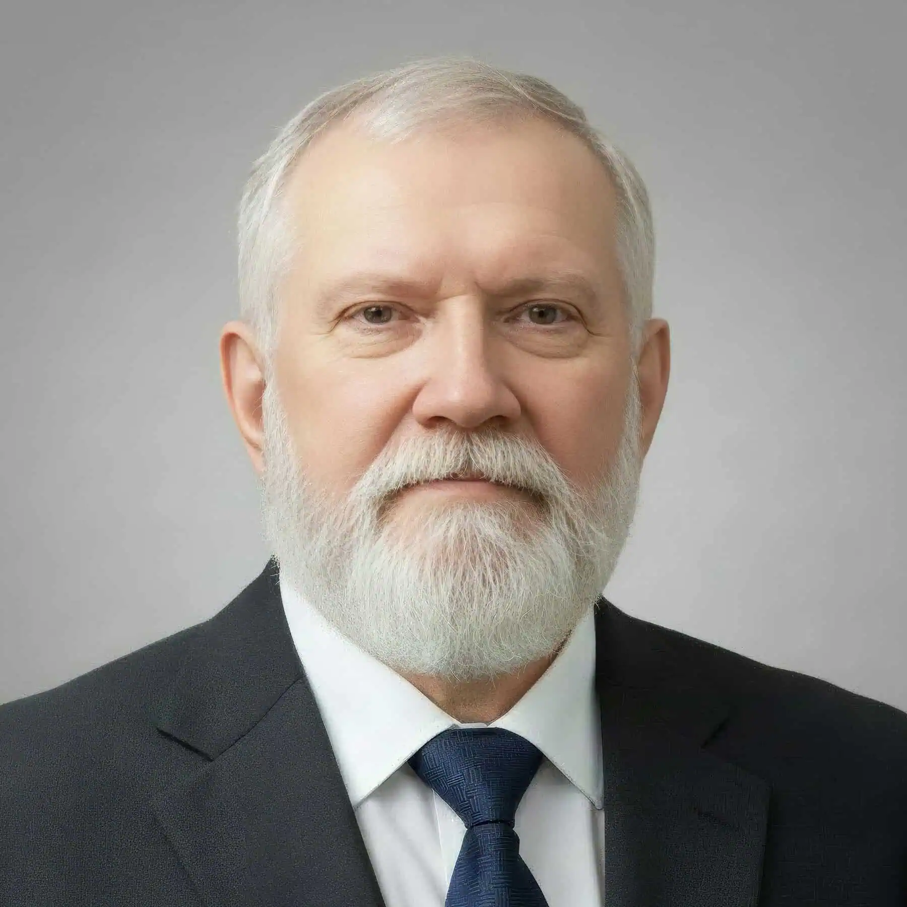 Photo of a white man with gray hair and beard wearing a dark suit, white shirt, and dark blue tie, with a gray background