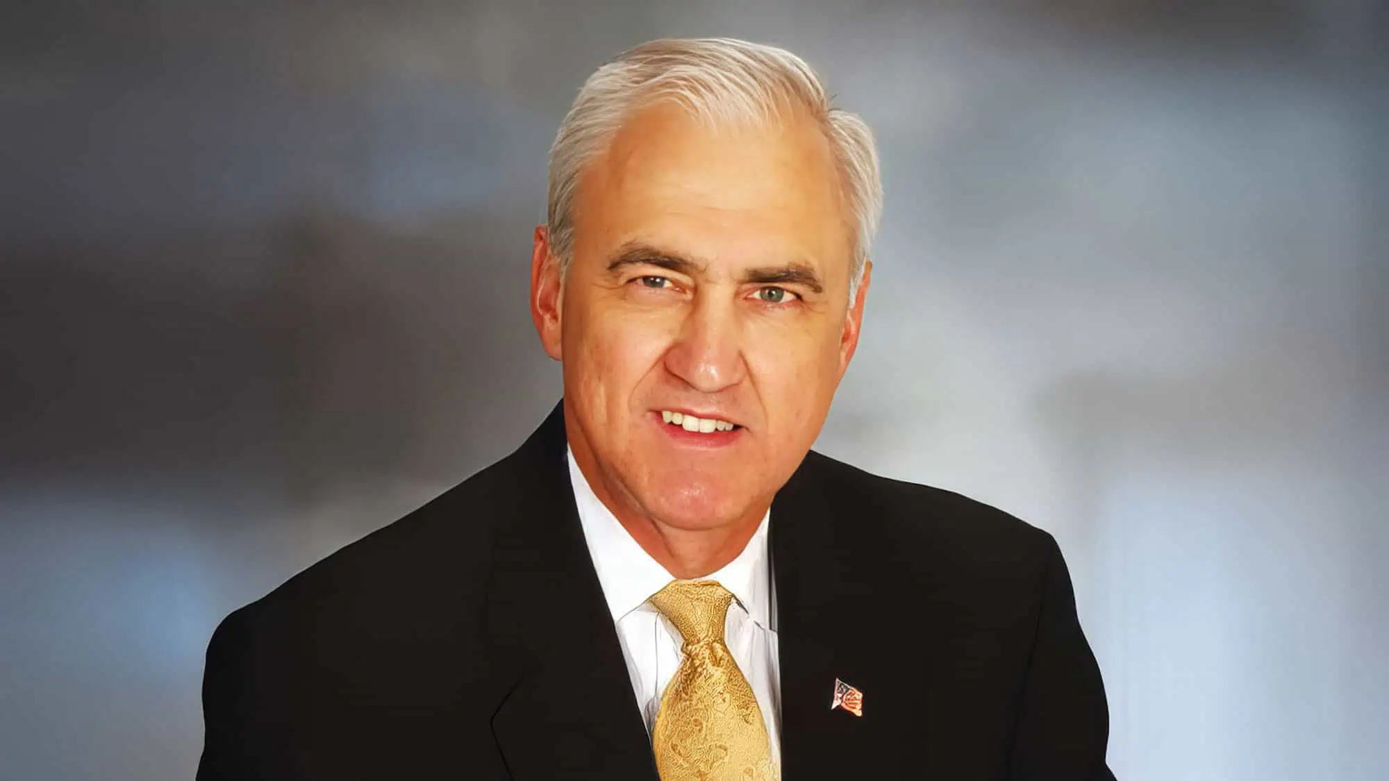 Photo of Jud Mason wearing a black suit, white shirt, gold, tie, and an American flag pin on his lapel with a formal photo portrait backdrop